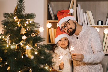 Family, winter holidays and people concept - happy bearded caucasian father and little cute daughter holding sparkles, bengal fire on background of decorating Christmas tree at home.