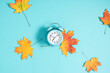 Alarm clock on blue background and autumn colorful leaves. Daylight saving time end. Fall leaves. Autumn background. Back to school concept. Flat lay.