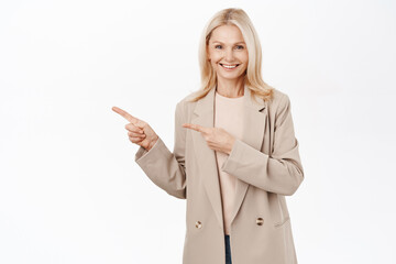 Portrait of smiling senior woman, 50 years old lady in trench coat, pointing finger left, looking happy and healthy, standing over white background