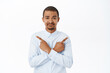 © Cookie Studio - Portrait of confused office worker, young man pointing sideways, cannot make decision, look clueless, standing over white background