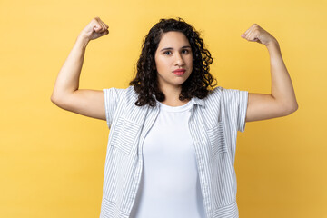 Yes, I am winner. Portrait of woman with dark wavy hair raises arms, demonstrates muscles, being powerful, ready to help you in difficult situation. Indoor studio shot isolated on yellow background.