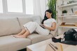 © SHOTPRIME STUDIO - Asian woman photographer lying at home on the couch and reviewing photos from the camera in laptop for color correction retouching, work as a freelance business photographer for yourself home office