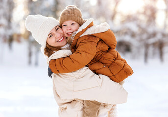 Happy family enjoying snowy weather outdoor, mom with little son hugging during walk in winter park