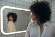 © Daniel - Young woman with dark curly hair checking clothes in the mirror in modern washroom.