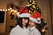 © sofiko14 - Portrait of happy mother and little cute daughter in santa hat and warm knitted white sweaters sitting near decorating tree in living room. Loving family indoors. Merry Christmas and Happy Holidays.