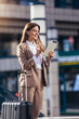 © Mediteraneo - Business woman travel to airport, hotel or office. Portrait of young professional walking in city street with suitcase bag using a digital tablet while waiting a driver
