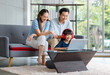 © Bangkok Click Studio - Millennial Asian happy family father and mother sit on cozy sofa couch smiling helping teaching little young girl daughter wearing headphones on floor studying learning online in living room at home