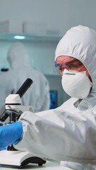 Wall Mural - Vertical video: Scientist in ppe suit placing a slide on specimen stage of a laboratory microscope making adjustments. Chemist in coverall working with various bacteria, tissue blood samples for