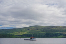 Luss, Scotland: A Small Boat On Loch Lomond, Mountains In The Distance, A Blue Sky With Thick White Clouds. Loch Lomond Is Part Of The Loch Lomond And The Trossachs National Park, Established In 2002.