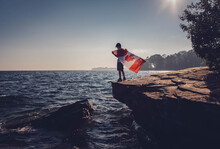 Full Length Of Boy Holding Canadian Flag While Standing On Cliff Against Sea And Sky During Sunny Day