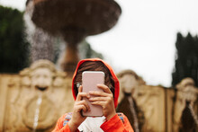 Boy Wearing Hooded Shirt While Using Smart Phone Against Fountain In City