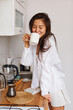 © olga pink - Asian brunette woman in white in kitchen. Female holding, drinking morning cup hot coffee tea