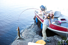 A Little Girl Holds A Fishing Pole With A Large Fish On The End Of The Fishing Line With The Boat In The Background.