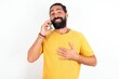 © Jihan - Smiling young bearded hispanic man wearing yellow T-shirt over white background  talks via cellphone, enjoys pleasant great conversation. People, technology, communication concept