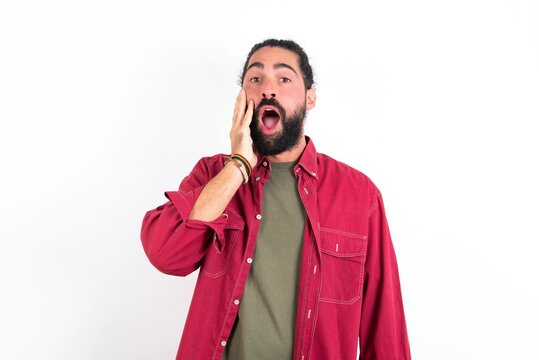 Shocked young bearded hispanic man wearing red overshirt over white background looks with great surprisment being very stunned, astonished with unexpected news, Facial expressions concept.