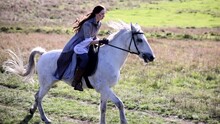 Young Woman In Image Of Ancient Warrior Rides On White Horse On Pathway Across Meadow.