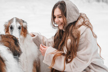 Young Beautiful Girl In Snowy Cold Winter Frost Forest Walking With Pet, Dog Of Hunting Breed Russian Borzoi. Sighthound, Wolfhound Owner. Having Fun, Laughing. Stylish Clothes, Fur Coat, Woolen Hat
