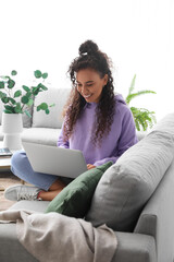 Wall Mural - Young African-American woman using laptop on sofa at home