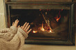 © sonyachny - Heating house in winter with wood burning stove. Woman in cozy sweater warming up hands at fireplace in rustic room. Young stylish female sitting at fireplace in farmhouse