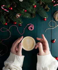 Female hands with white sweater sleeves hold a cup of coffee on a green wooden table in a Christmas decor