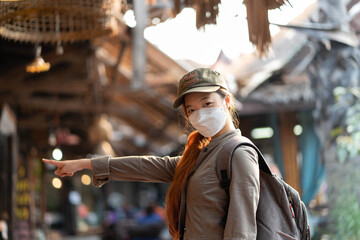 Wall Mural - Young asian woman traveler with mask and backpack pointing finger at community of thailand