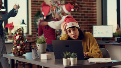 Wall Mural - Festive people disturbing stressed woman during christmas time, feeling frustrated by noisy colleagues. Coworkers celebrating winter holiday season, interrupting tired person. Tripod shot.