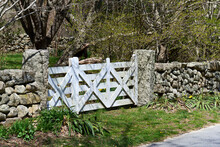 A White Wood Cross-braced Gate Is Set In A Stone Wall Alongside A Road.