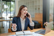 © amnaj - Beautiful Asian business woman chatting on the phone with customer and working on her laptop and taking note enthusiastically and happily in the office.