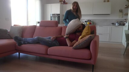 cheerful playful young caucasian couple having pillow fight in modern cozy living room at home. happ
