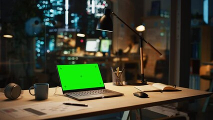 Fototapete - Laptop Computer Standing on a Desk with a Green Screen Chromakey Mock Up Display in Modern Creative Office at Night. Employee Working in Warm Evening Lighting In the Background.