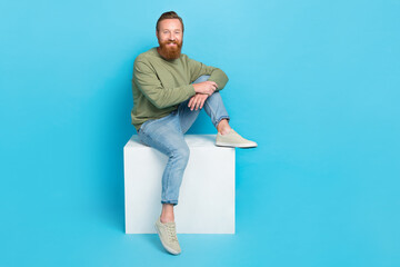 Wall Mural - Full size photo of satisfied pleasant man with red hairstyle khaki pullover sitting on white cube box isolated on blue color background