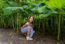 Contented Girl In A Dress With Blue Stripes Is Sitting Under Huge Rhubarb Leaves Outdoors
