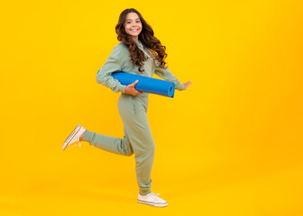 Poster - Full length of a fitness teen girl in sportswear hold yoga mat posing over yellow background. Fitness model child wearing sport clothes. Run and jump. Girl in the sport concept.