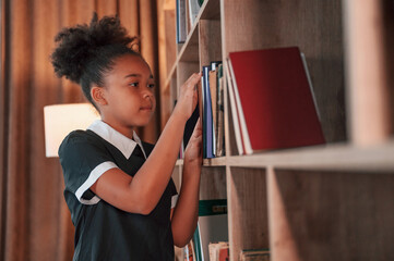 Wall Mural - Searching for the books on the shelves. Cute african american girl in school uniform is at home library