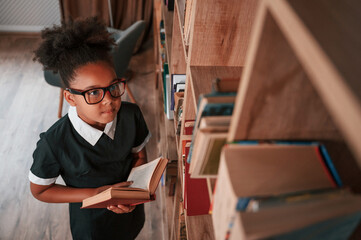 Wall Mural - Searching for the books on the shelves. Cute african american girl in school uniform is at home library