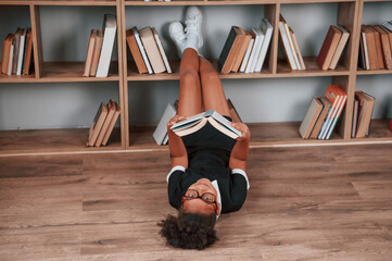 Wall Mural - Laying down on the floor and reading the book. Cute african american girl in school uniform is at home library