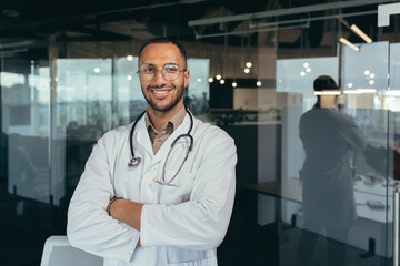 Portrait of a young handsome male hispanic doctor. Standing in the glass hall of the hospital wearing glasses, a white coat and a stethoscope. He looks at the camera, crosses his arms, smiles.