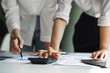 © crizzystudio - Working as a team with analytical graphs, business people are using calculators to calculate investment, turnover and profits displayed in a document on a desk at a conference room.
