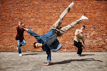 Freeze Frame Of Male Breakdance Performer Doing Handstand Pose With Team Against Brick Wall Outdoors