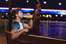 Handsome Sporty Man Drinking Lemonade After Workout, Sitting On A Bench On The Riverside