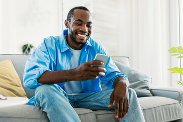 Wall Mural - Cool gadget and app concept. Portrait of happy black man using mobile phone, watching video, sitting on sofa copy space