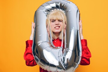 a woman in a stylish red shirt stands on a yellow background and making a funny face expressing emotions holds a balloon in the form of the number zero in her hands, sticking her head into it