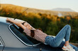 canvas print picture - The woman driver stopped on the road and lay down on the car to rest and look at the beautiful landscape in a striped T-shirt and jeans. Complicated journey to nature