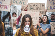 © MandriaPix - People strike against climate change and pollution, portrait of young woman holding a megaphone and shouting, green new deal protest, dark mood filter