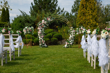 Poster - A wedding arch of many flowers on a green lawn in a park against the backdrop of decorated chairs, coniferous bushes and trees