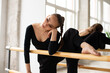 Modern female dancer in black outfit standing in front the mirror leaning on wooden handrail while practices in a dance studio