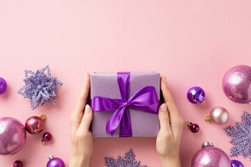 Christmas concept. First person top view photo of female hands giving lilac giftbox with ribbon bow over violet baubles snowflake and flower ornaments on isolated pastel pink background with copyspace