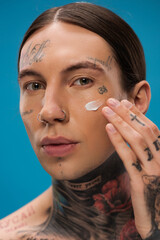 tattooed young man applying face cream on cheek and looking at camera isolated on blue