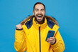 © ViDi Studio - Young happy man wear yellow down jacket hold in hand use mobile cell phone surfing internet do winner gesture isolated on plain blue color background studio portrait. People winter lifestyle concept