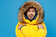 Young frozen sad caucasian man 20s wearing yellow puffer down jacket with fur hood hug himself try to warm up isolated on plain blue color background studio portrait. People winter lifestyle concept.